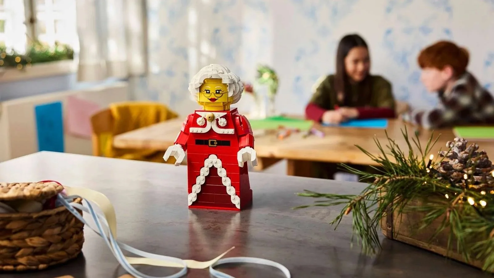 LEGO Up-Scaled Mrs. Claus Minifigure in a red dress with white trim standing on a kitchen counter, with children crafting behind her