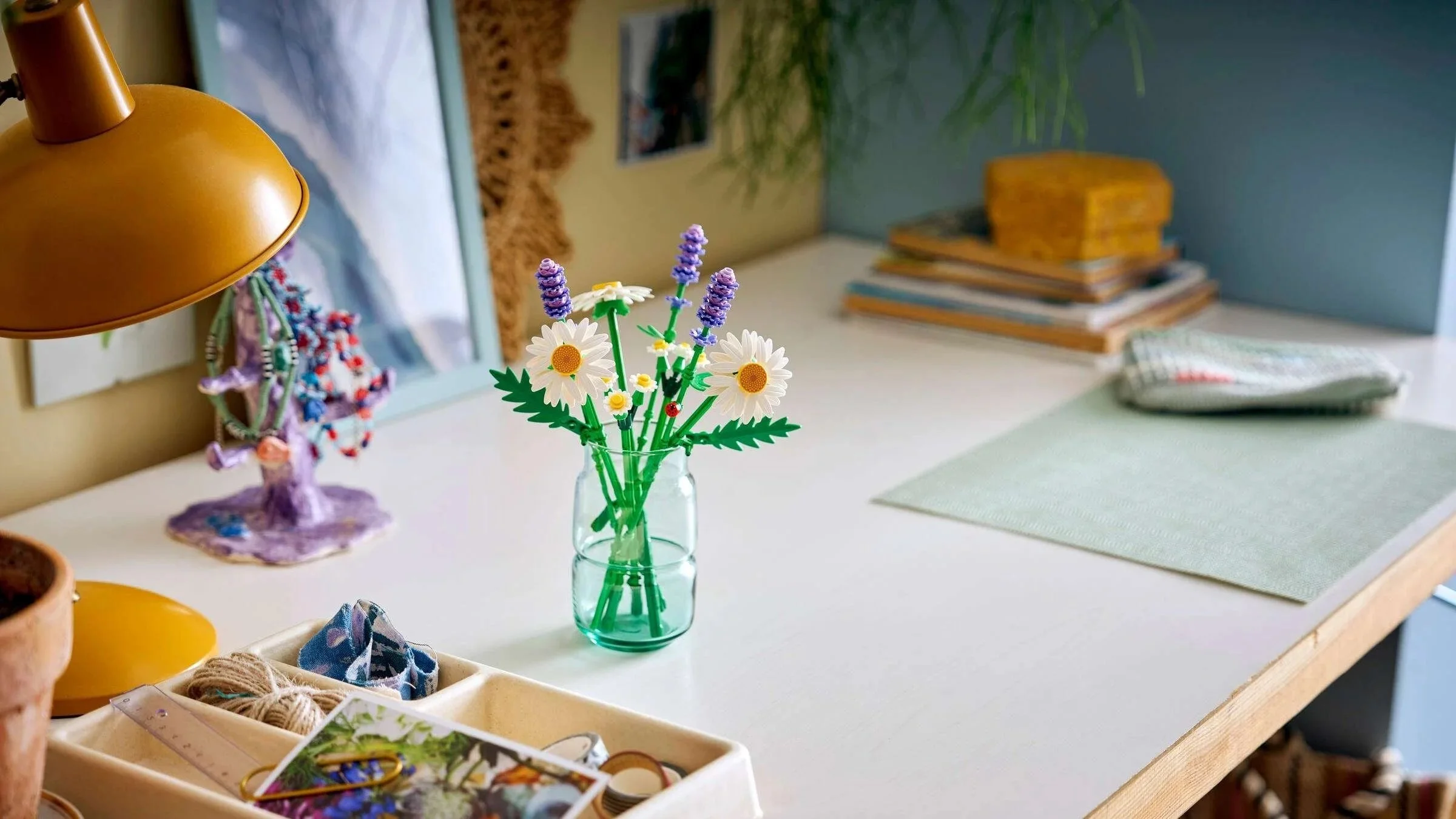 LEGO Daisies set with white daisies and lavender stems in a green glass vase on a white desk beside a mustard lamp