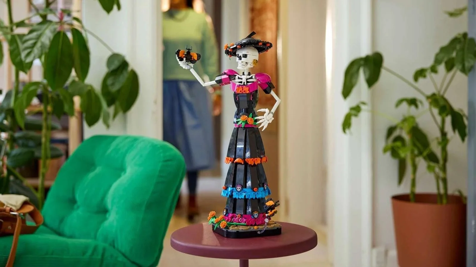A LEGO La Catrina figure raises a small skeleton dog on an outstretched arm, displayed on a round table in a plant-lined hallway