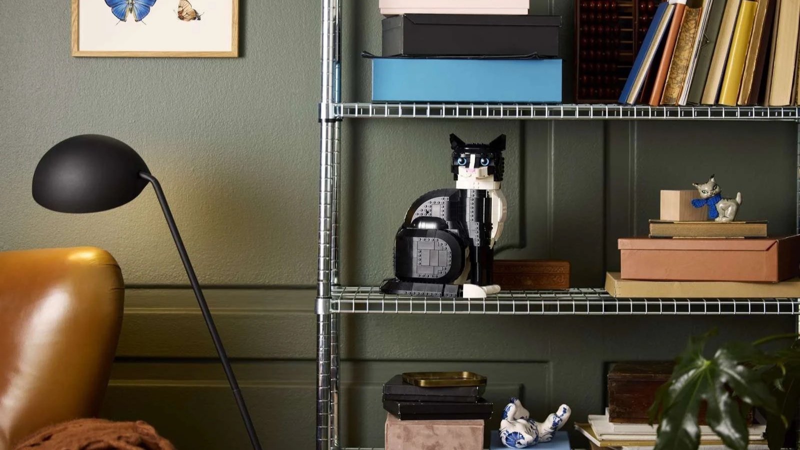 Black and white LEGO tuxedo cat figure on a wire shelf, with a butterfly art print on the wall behind
