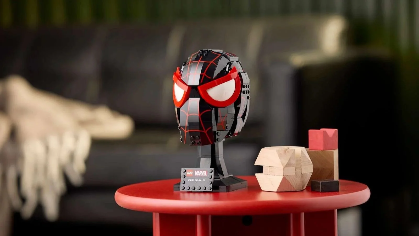 LEGO Marvel Miles Morales' Mask on a small red side table beside wooden puzzle blocks, with a sofa in the background