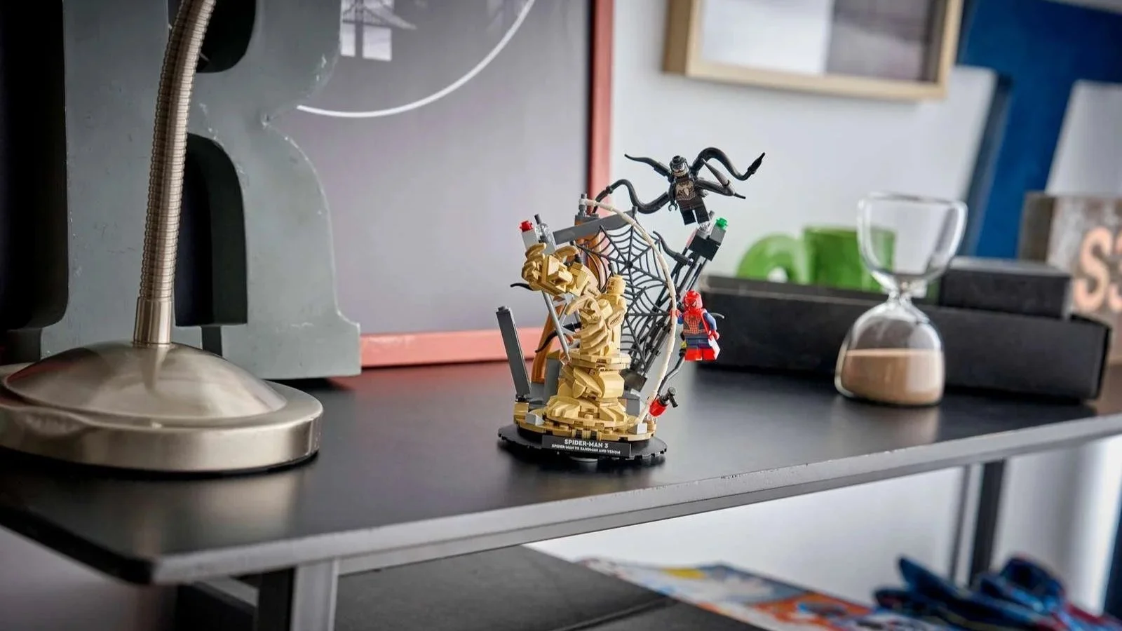 LEGO Epic Battle Spider-Man vs. Sandman display set with Spider-Man, Sandman and Venom around a web, on a black desk shelf next to a lamp and hourglass