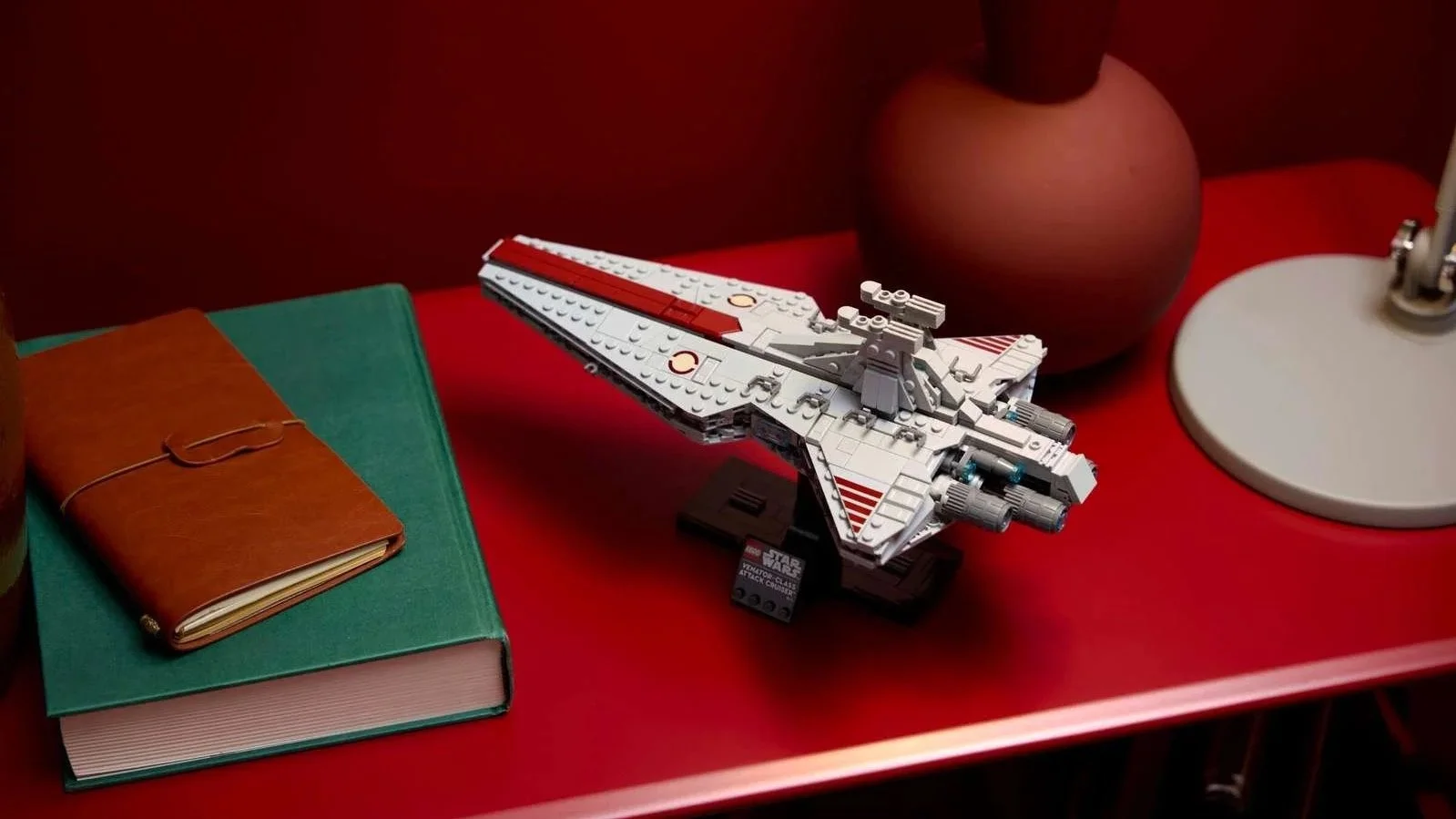 White and red LEGO Venator-Class Attack Cruiser on a black display stand, set on a red table next to a green book and a leather notebook