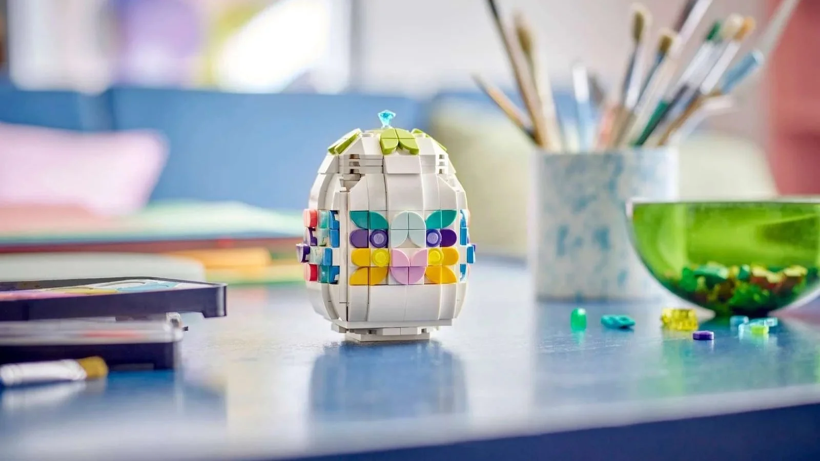 LEGO Decorative Easter Egg set, a white egg built from bricks with pastel flower patterns, on a blue table next to a cup of paintbrushes
