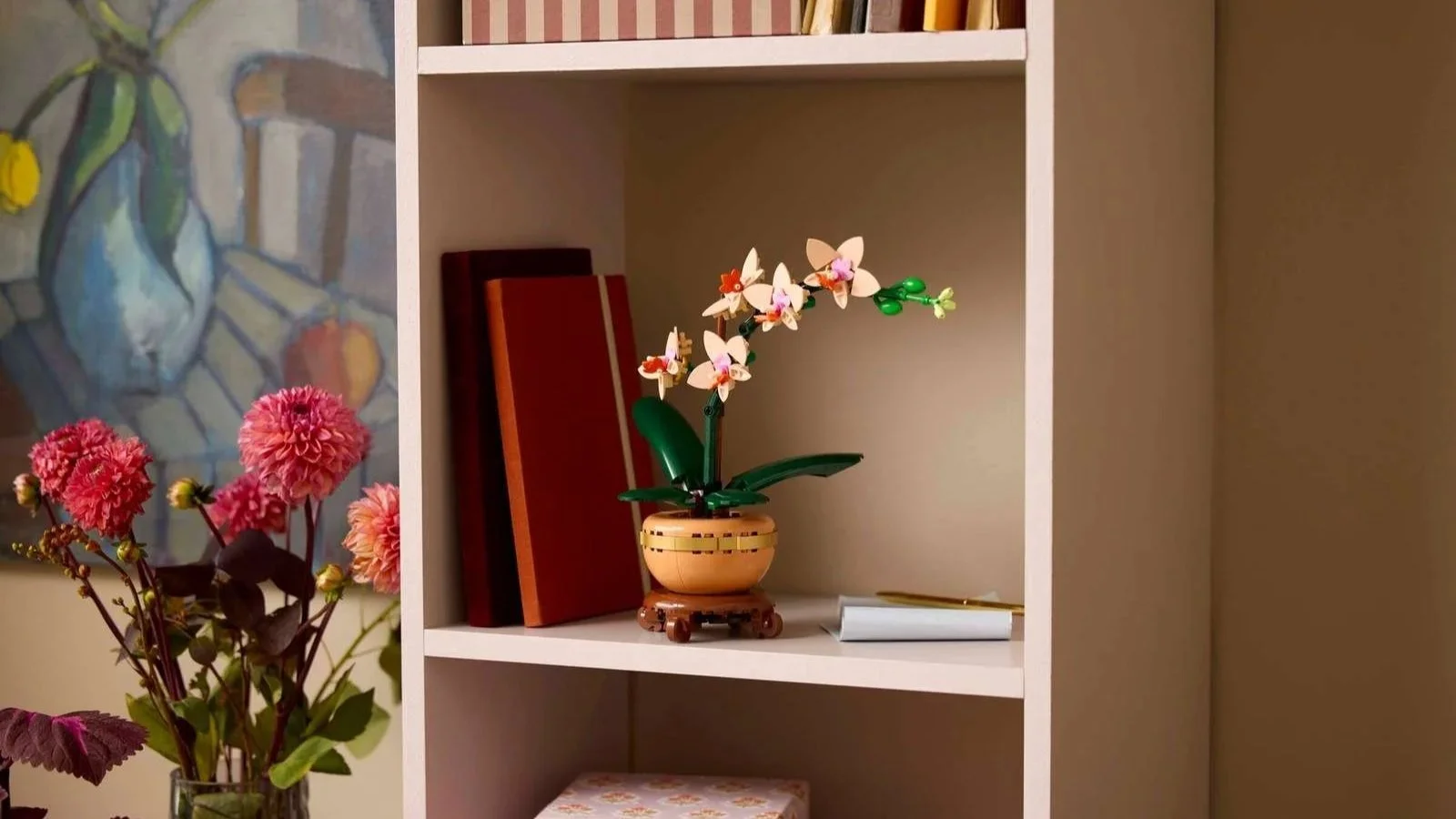 A LEGO Mini Orchid with pink and white blooms sits on a shelf beside stacked books and a framed botanical painting