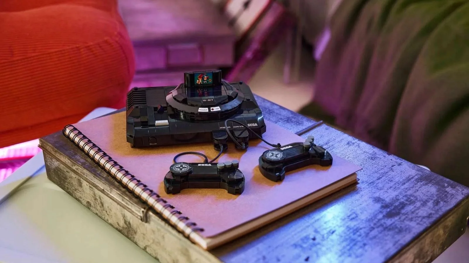 LEGO SEGA Genesis Console set with two controllers and a game cartridge, resting on a notebook beside a red armchair