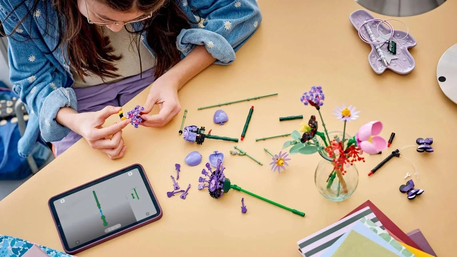Woman building purple LEGO flowers at her desk, with a finished bouquet in a vase and tablet instructions