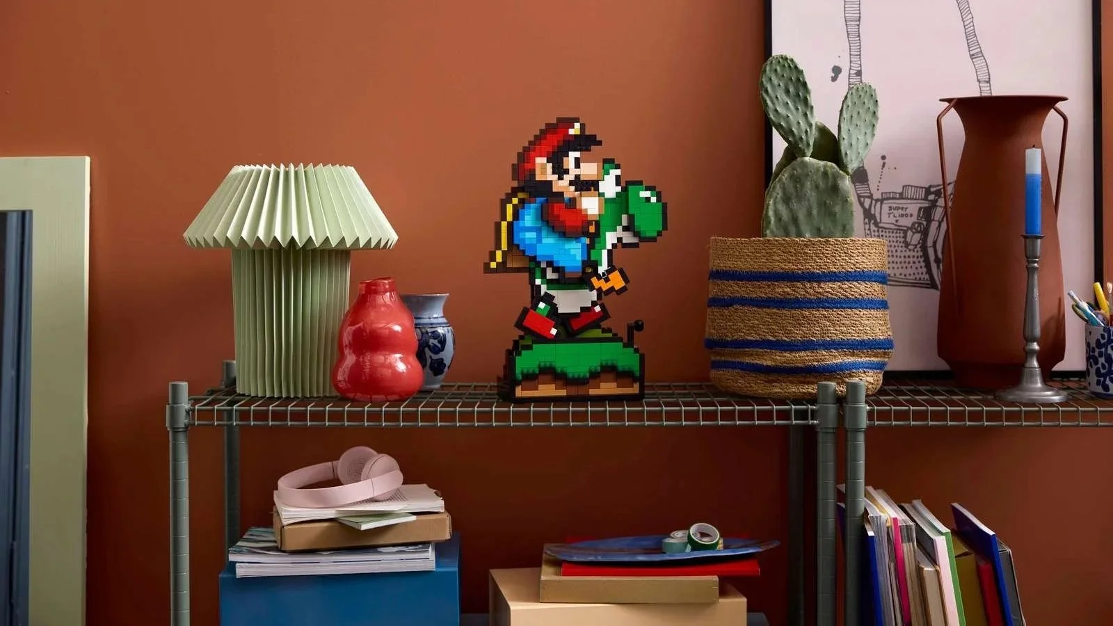 Pixel-art Mario rides Yoshi in this LEGO Super Mario World build, displayed on a wire shelf between a potted cactus and a line-art print