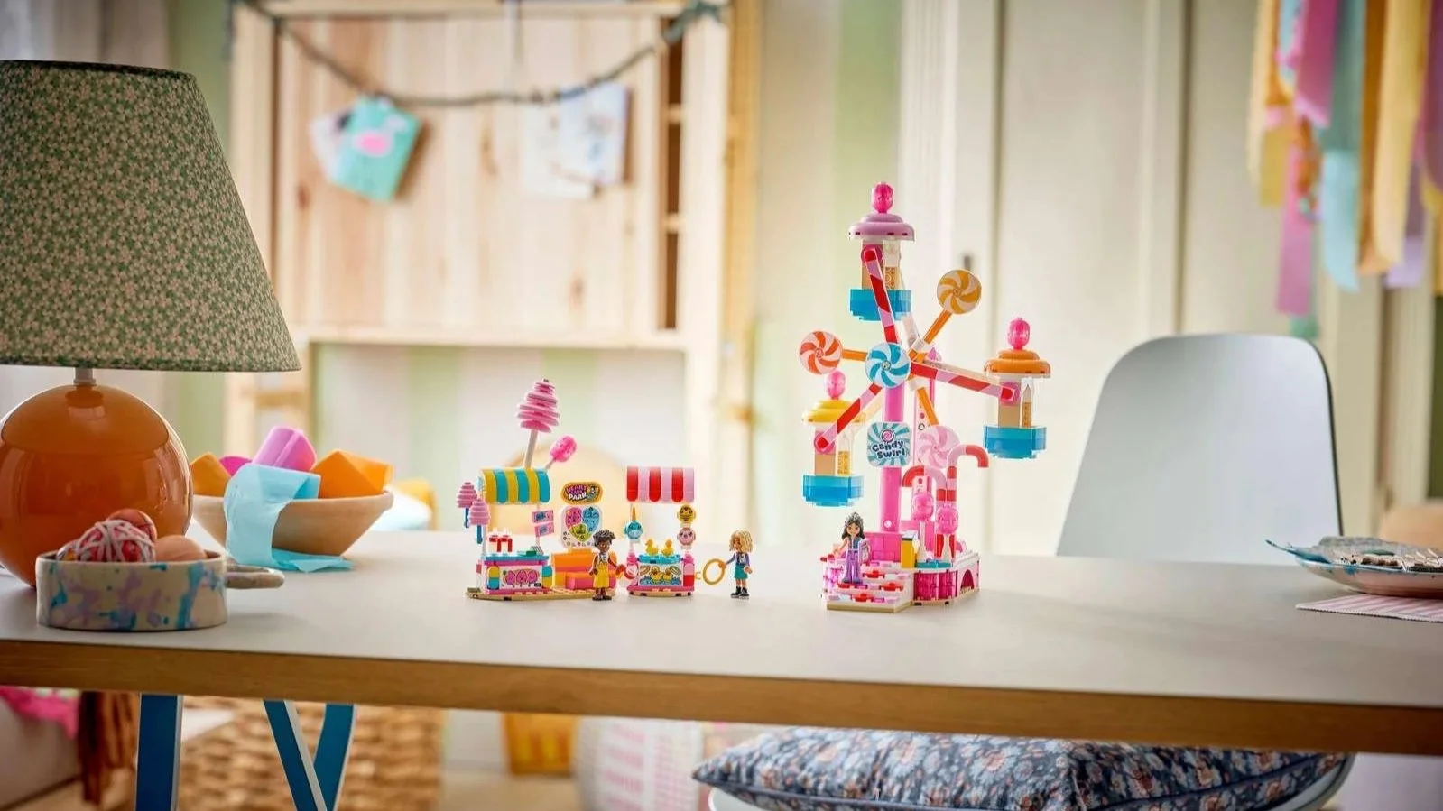 LEGO Candy and Cupcake Ferris Wheel set on a desk, a pink ferris wheel with lollipop spokes next to two striped-awning carnival stalls and three figures