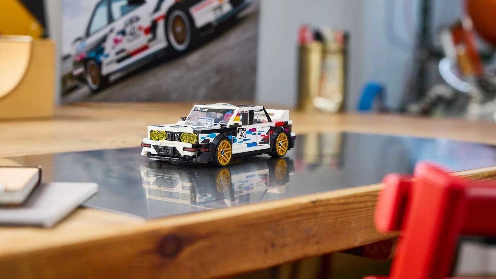 LEGO BMW M3 race car model in white and black with colorful racing graphics, number 40, and gold wheels, displayed on a desk in front of a matching poster