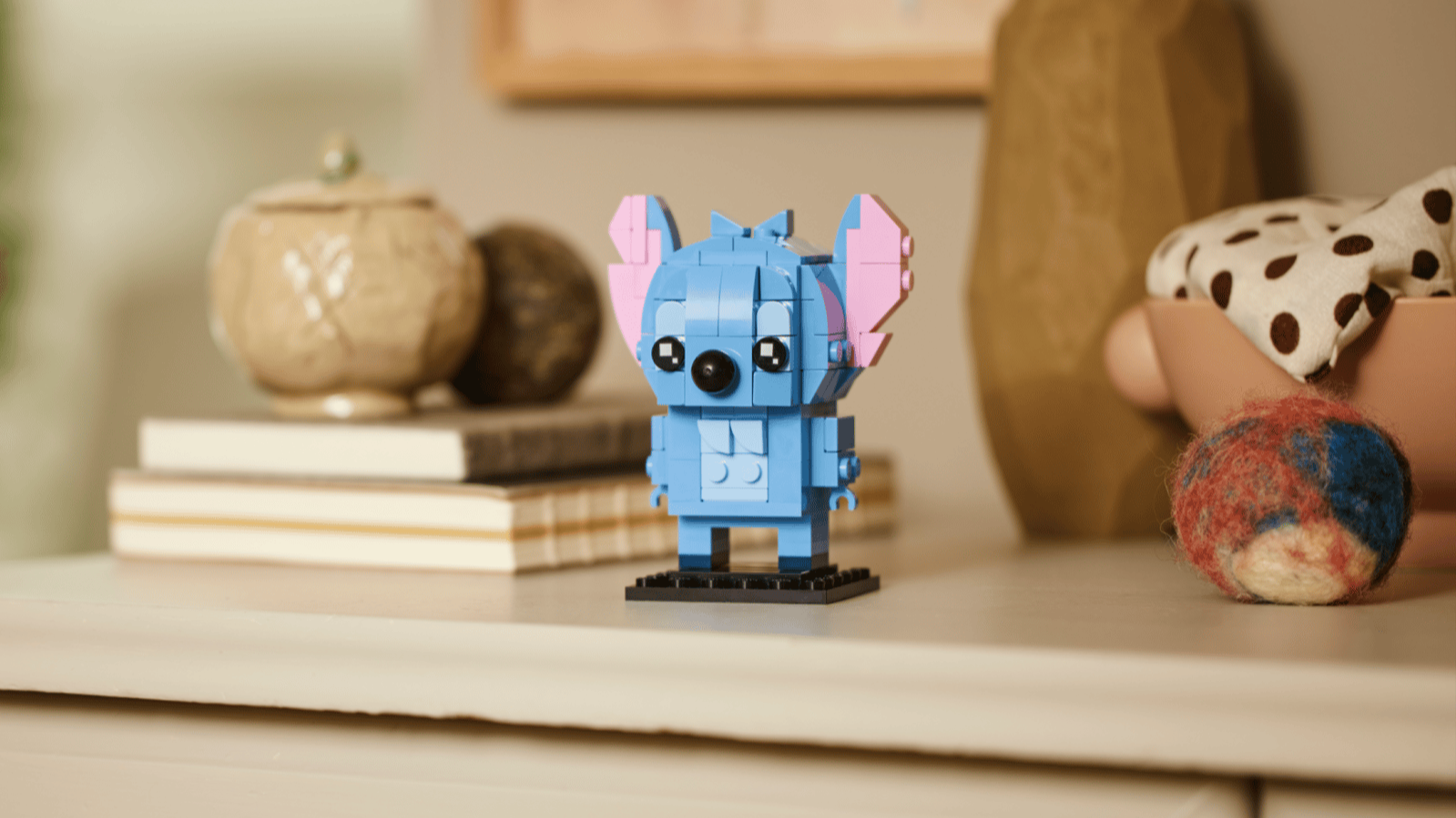Blue LEGO Stitch figure with pink ears on a black base, displayed on a cream dresser beside stacked books and a ceramic jar