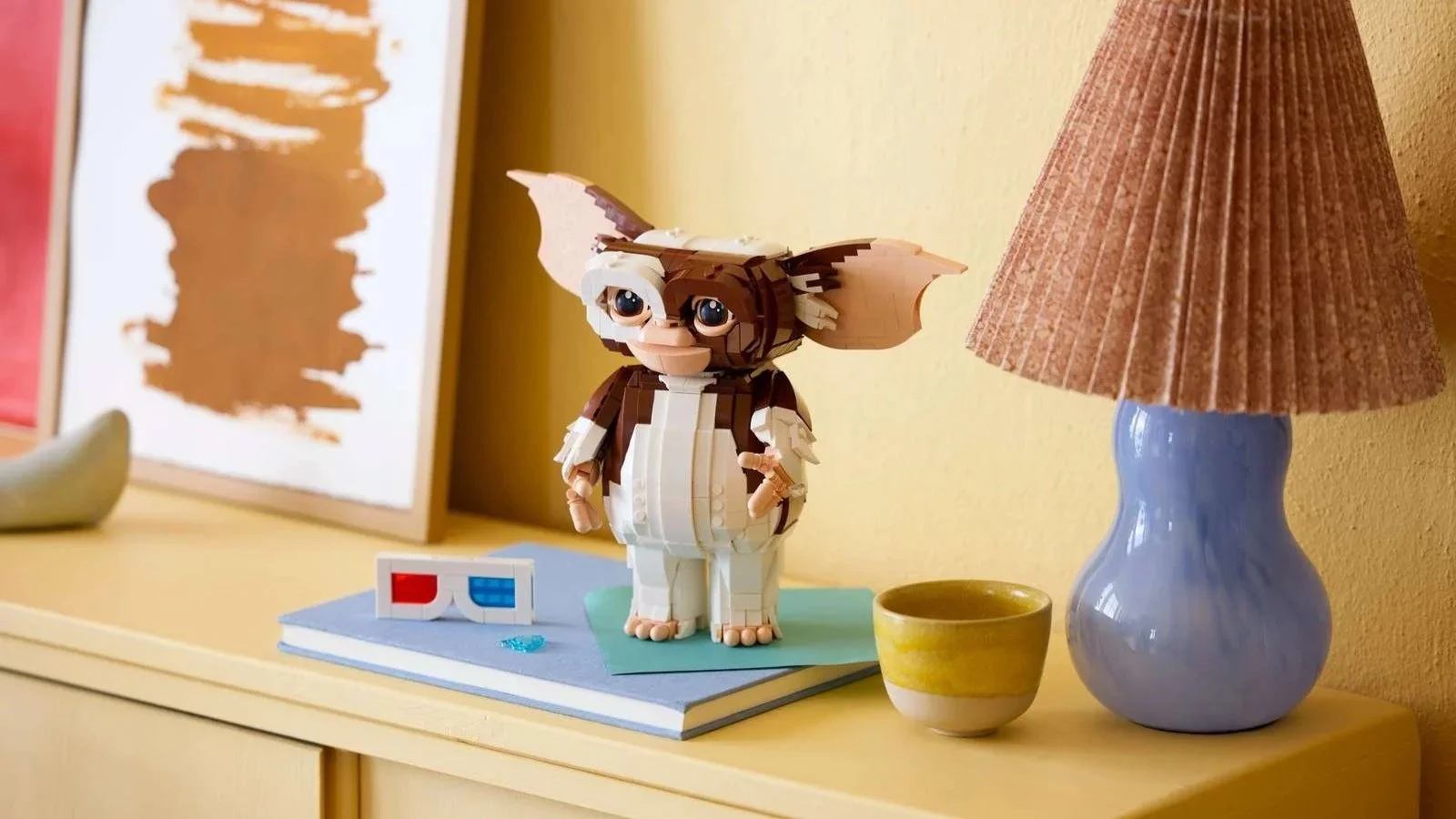 The LEGO Gizmo set from Gremlins standing on a yellow sideboard, with brick-built 3D glasses resting on a book beside it.