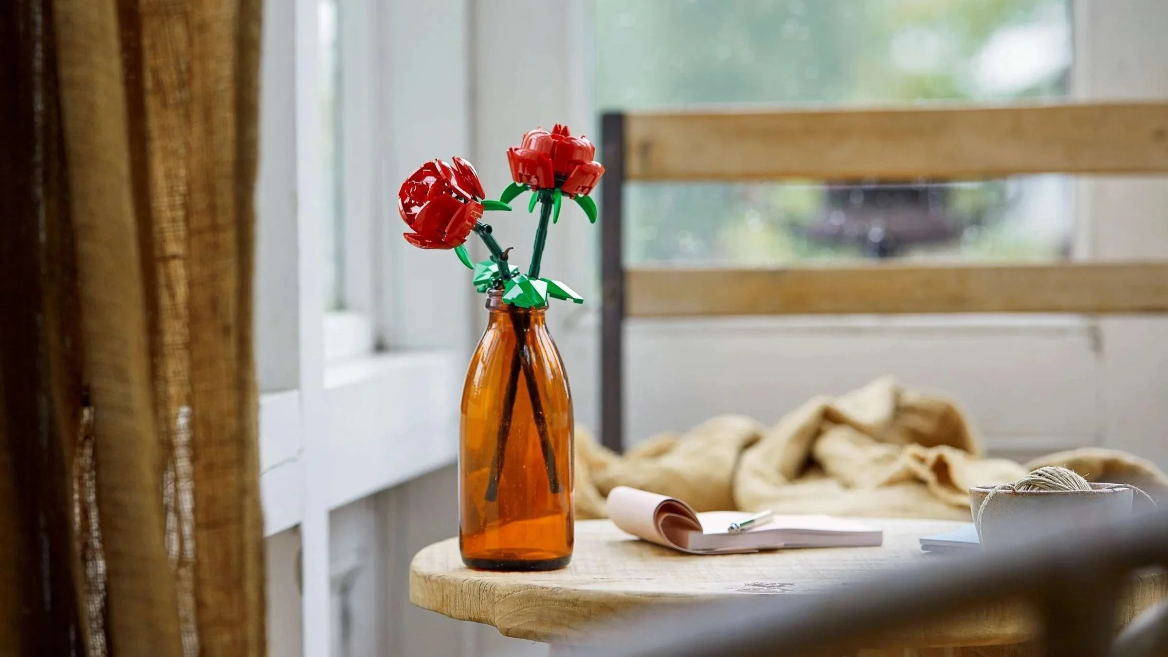 Red LEGO Roses in an amber glass bottle on a small wooden table beside a rainy window