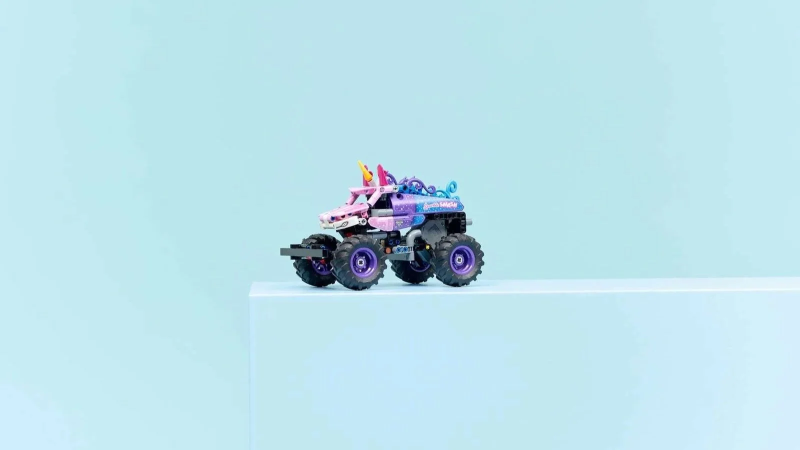 LEGO Monster Jam Sparkle Smash truck styled like a unicorn, pink and purple with a horn and flowing mane, perched on the edge of a light blue block