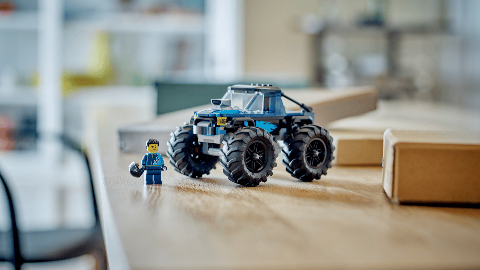 LEGO Blue Monster Truck set with oversized black tires and blue flame decals beside a minifigure holding a helmet on a wooden desk