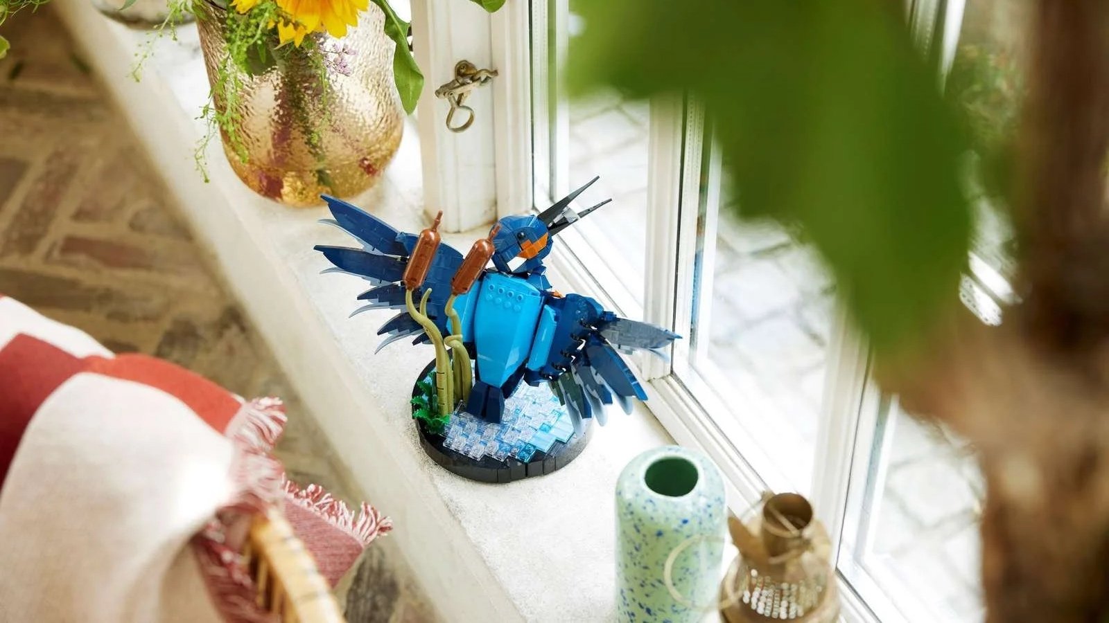 A LEGO Kingfisher Bird sculpture perched on a stone windowsill beside a vase of sunflowers, with a courtyard visible outside