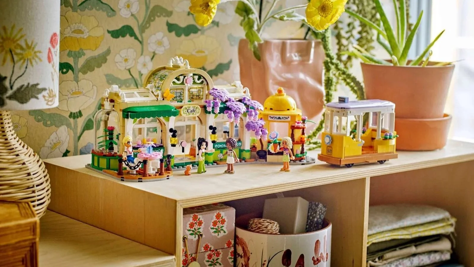 LEGO Friends Plant Café and Flower Shop set on a wooden shelf, a glass-roofed café with purple wisteria, a yellow flower kiosk, and a small yellow tram