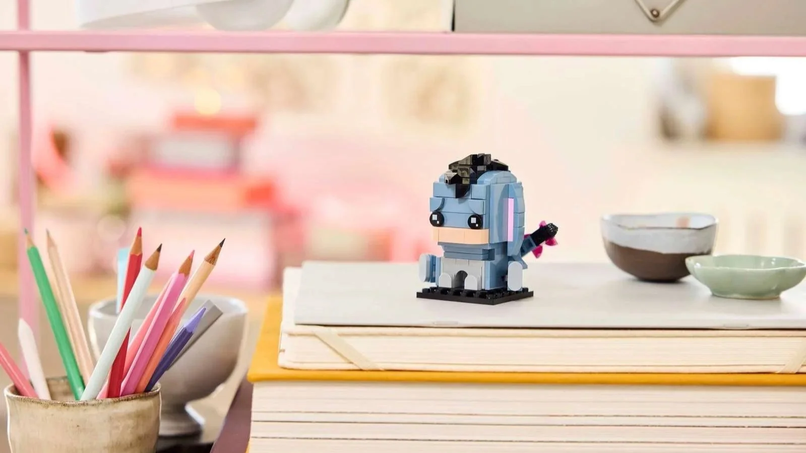 LEGO Eeyore figure with a blue-gray blocky head and pink bow on its tail, sitting on a stack of notebooks next to a cup of colored pencils