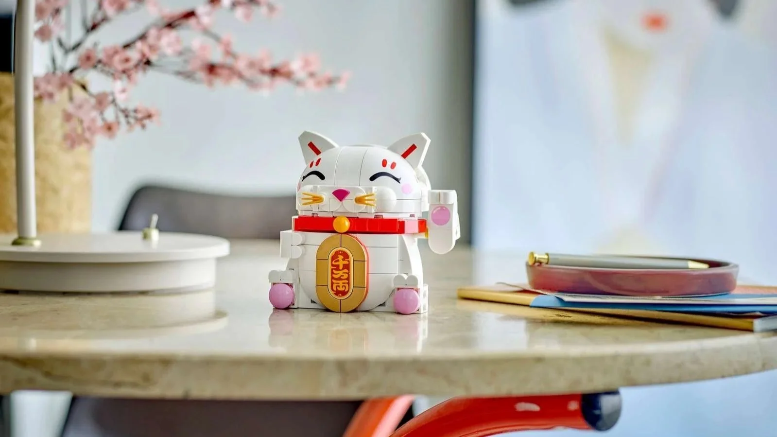 White LEGO lucky cat with a raised paw, red collar and gold coin on a table beside cherry blossom branches