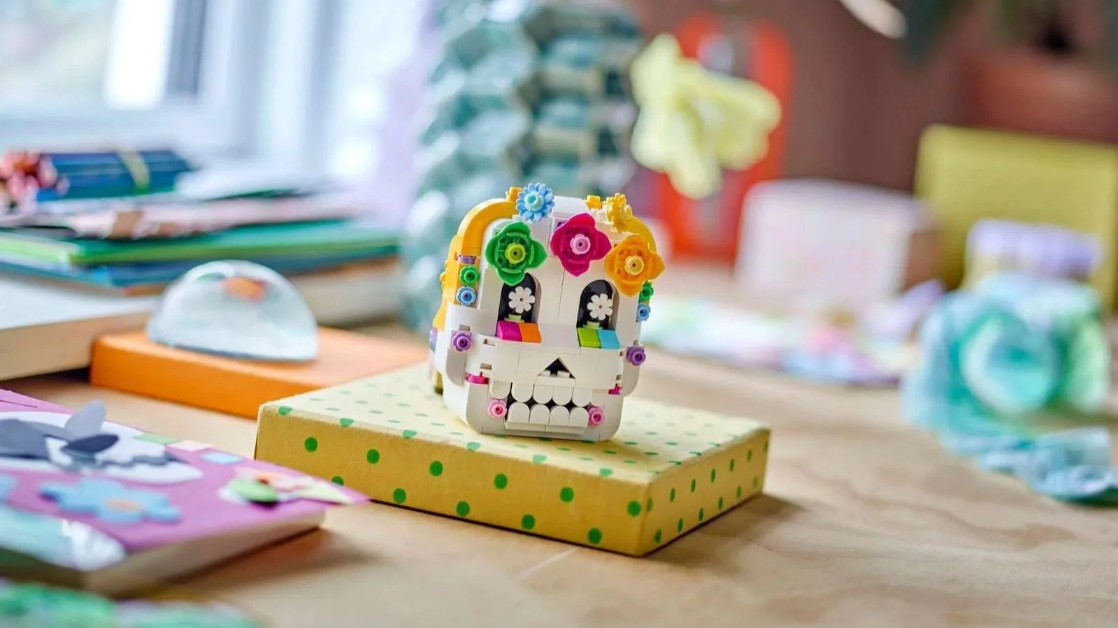 LEGO Altar of the Dead set, a white sugar skull with colorful brick flowers, displayed on a polka dot gift box on a wooden desk