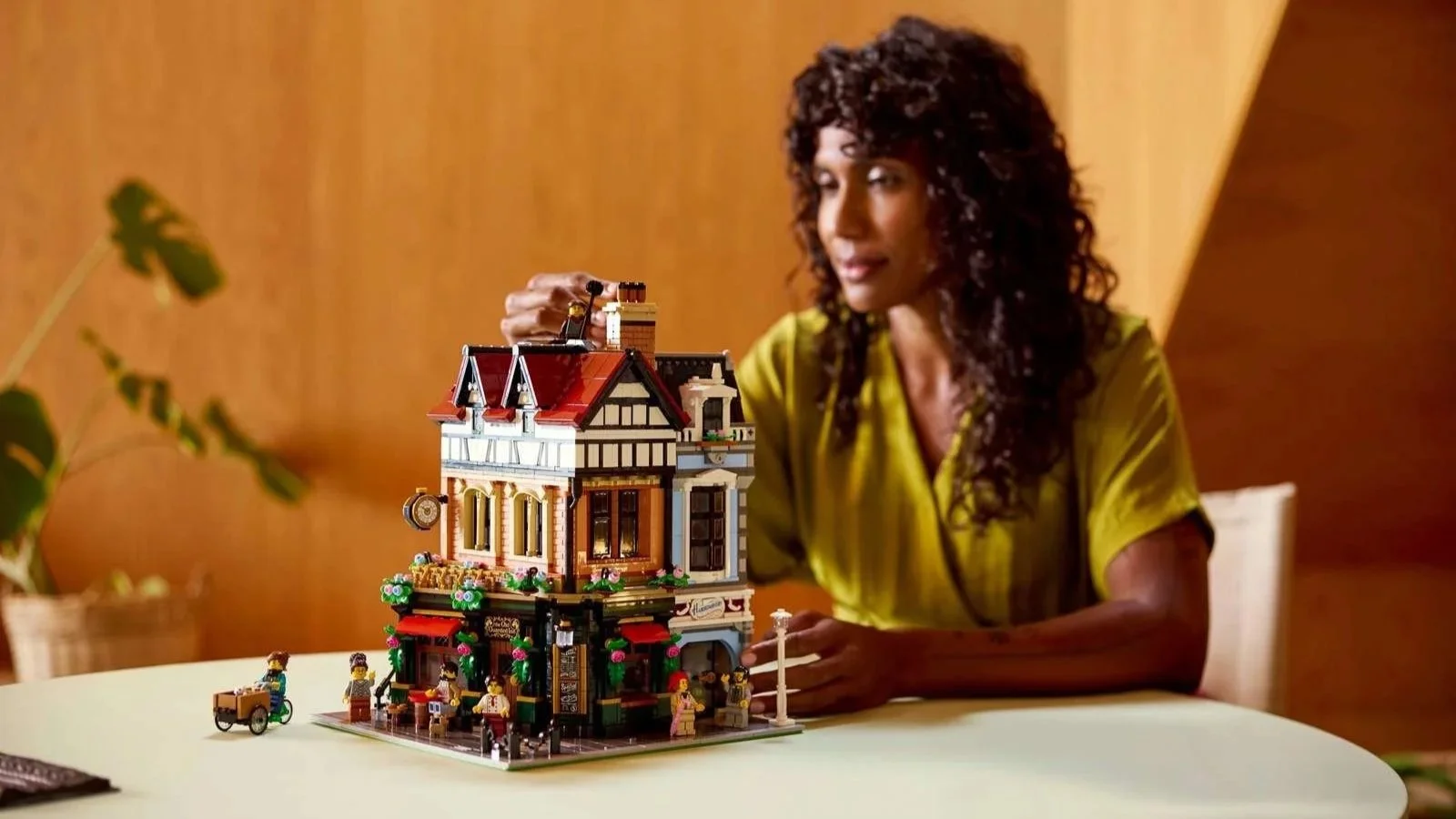 A woman placing a piece on the roof of a LEGO Tudor Corner building with its red tiled gables, flower boxes, and a tiny cargo bike parked out front