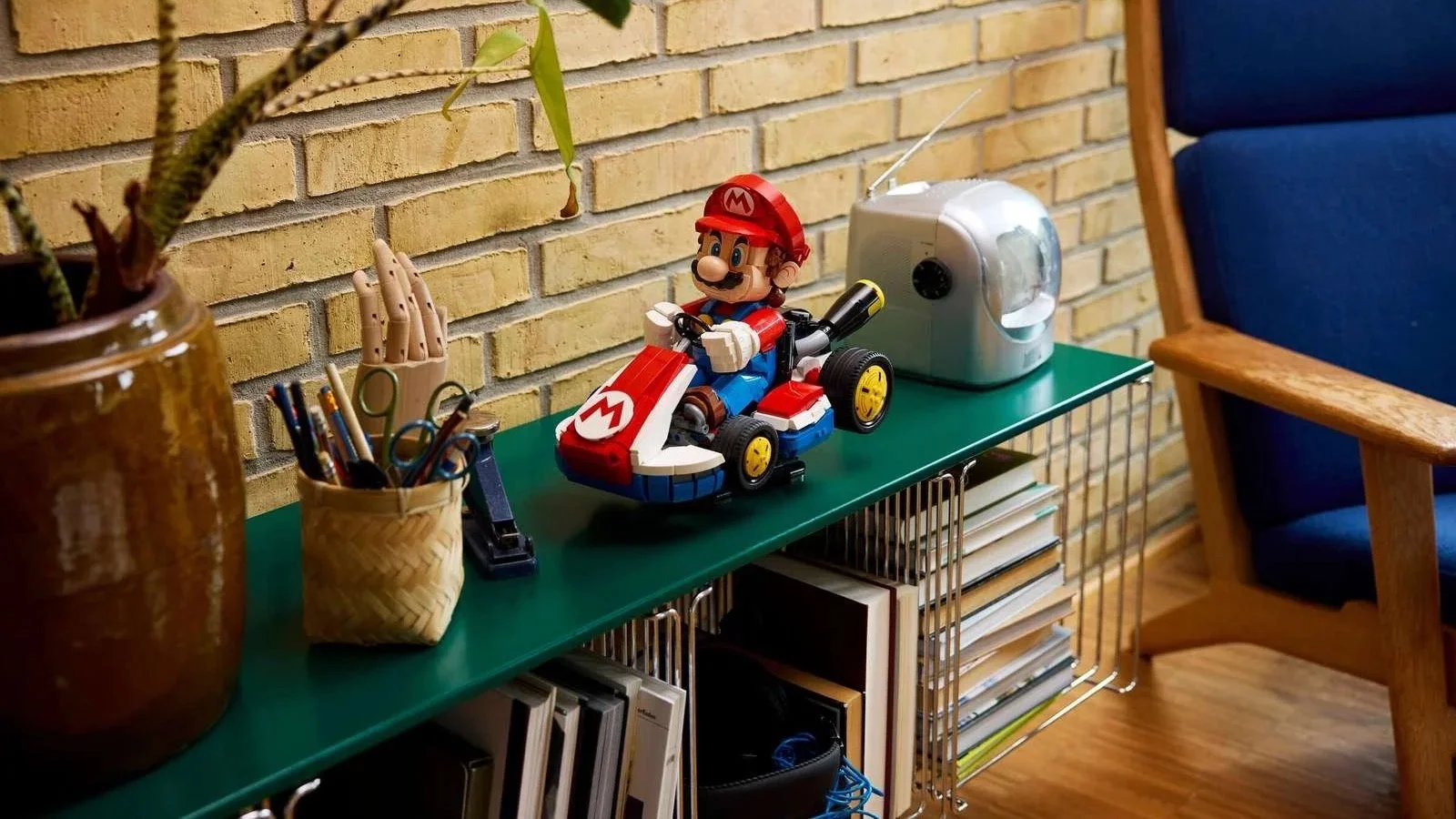 A LEGO Mario figure races his Standard Kart across a green console table, flanked by a potted plant and a vintage radio