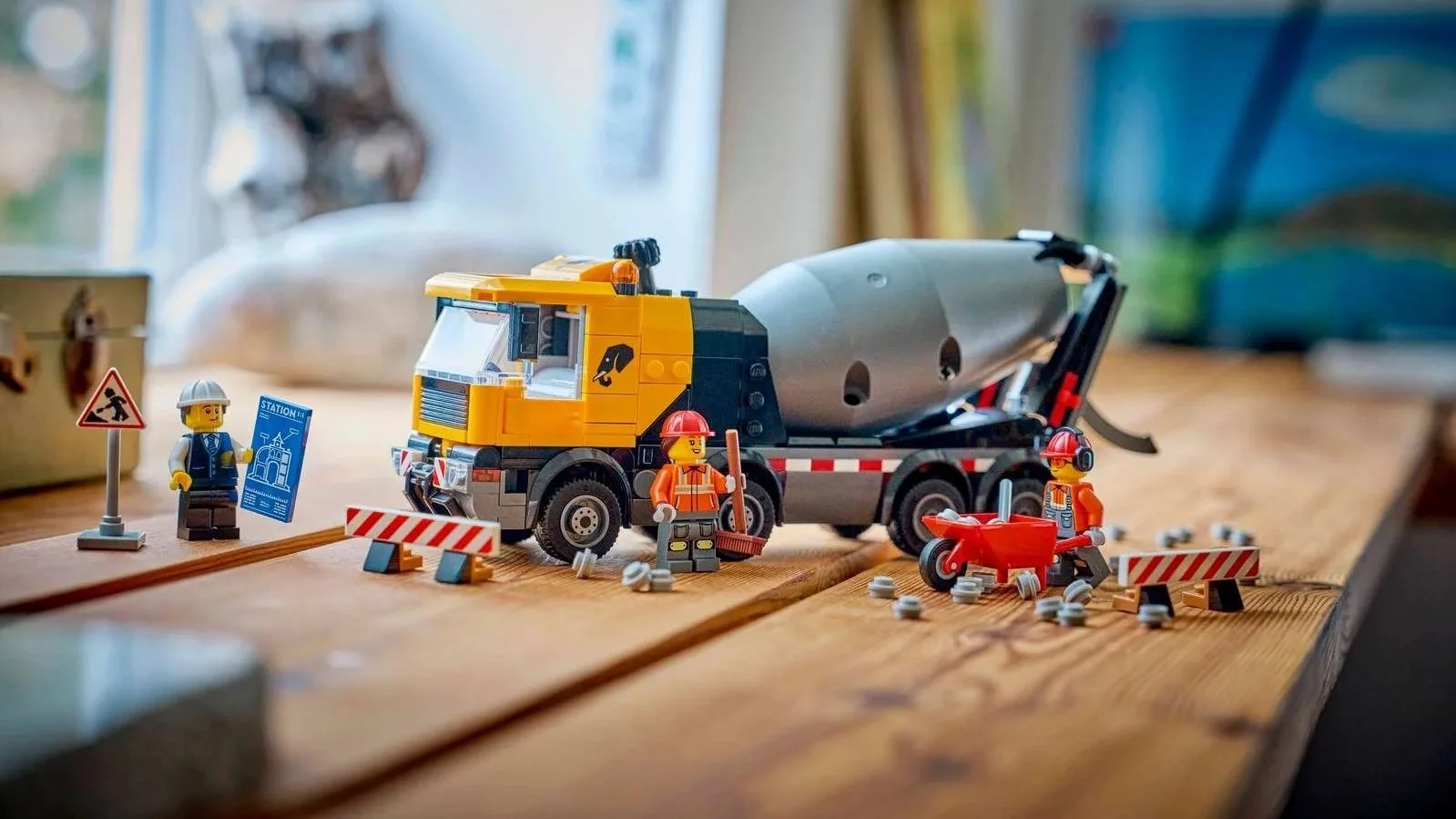 LEGO Cement Mixer set with a yellow cab and gray drum, three construction minifigures, road barriers and a red wheelbarrow on a wooden desk
