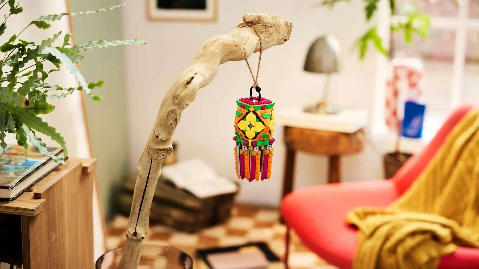 A LEGO Colorful Lantern hangs from a driftwood branch above a glass vase in a plant-filled living room
