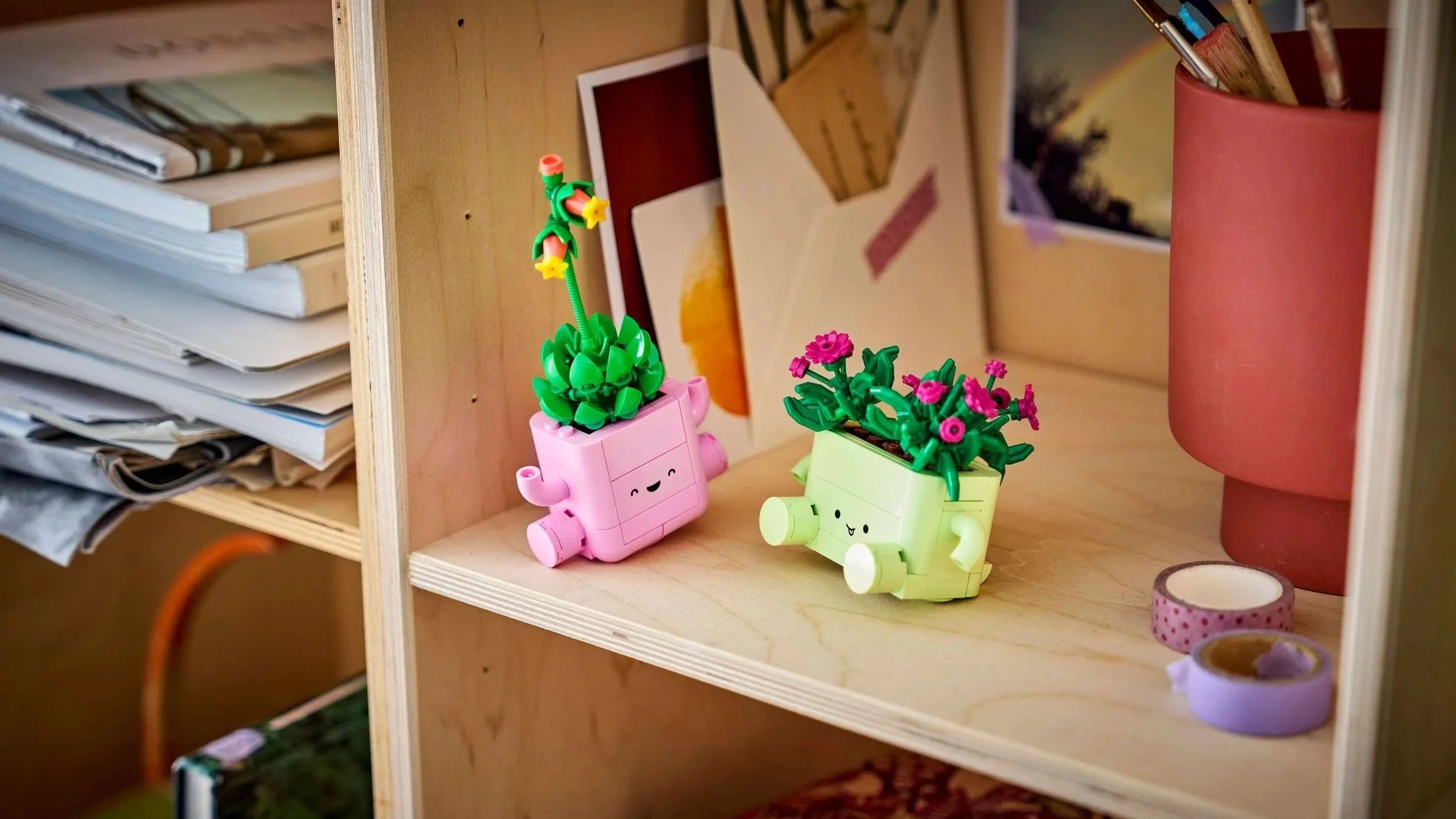 Two LEGO Rocking Plants pots with smiling faces, one holding a succulent and one holding pink flowers, on a wooden shelf