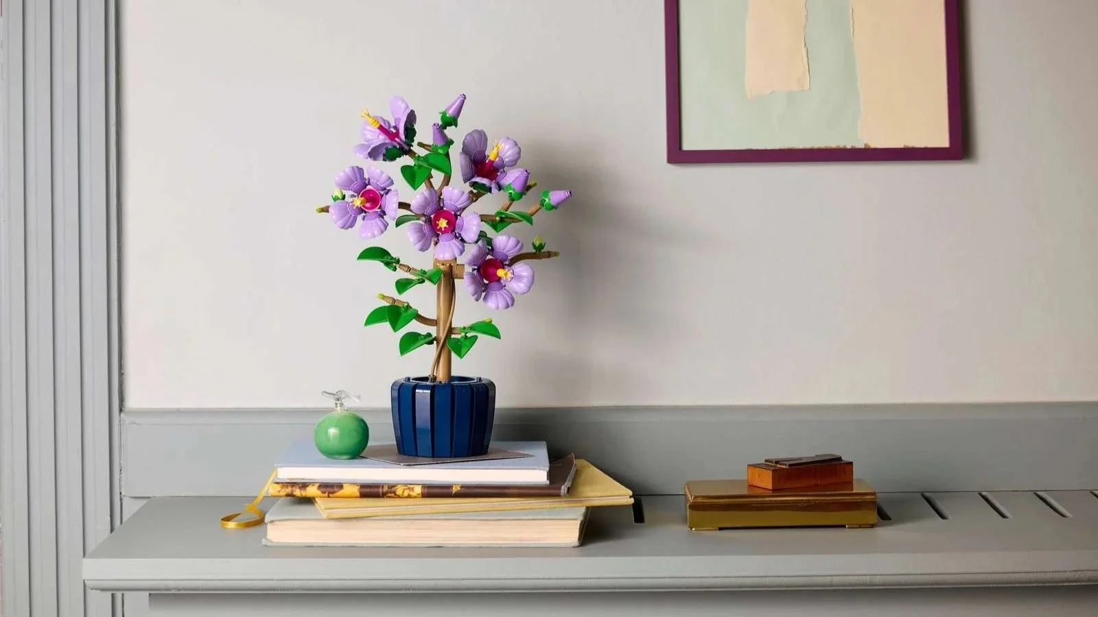 LEGO Hibiscus set with lavender blooms in a dark blue pot, displayed on a stack of books on a gray shelf
