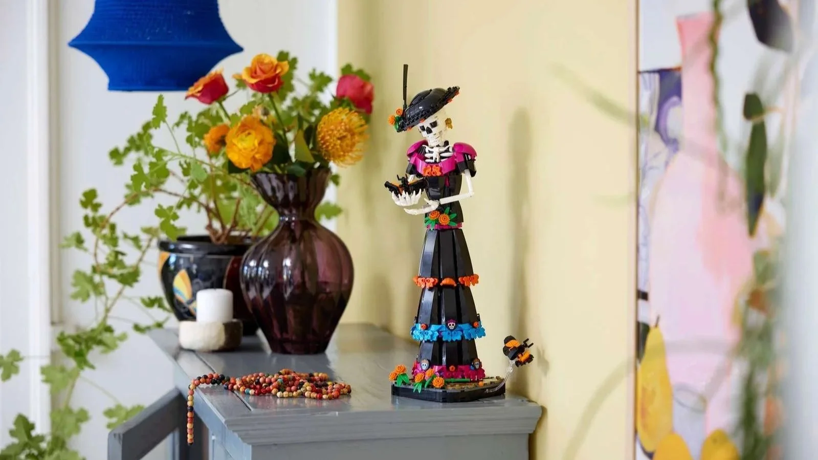 A LEGO La Catrina figure in a black and pink gown stands on a cabinet beside vases of fresh flowers and a strand of beads