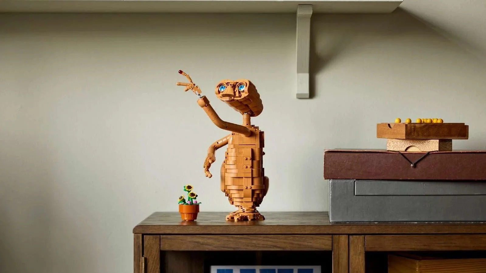 LEGO E.T. the Extra-Terrestrial figure with one arm raised, standing beside a small LEGO sunflower pot on a wood cabinet below a floating shelf
