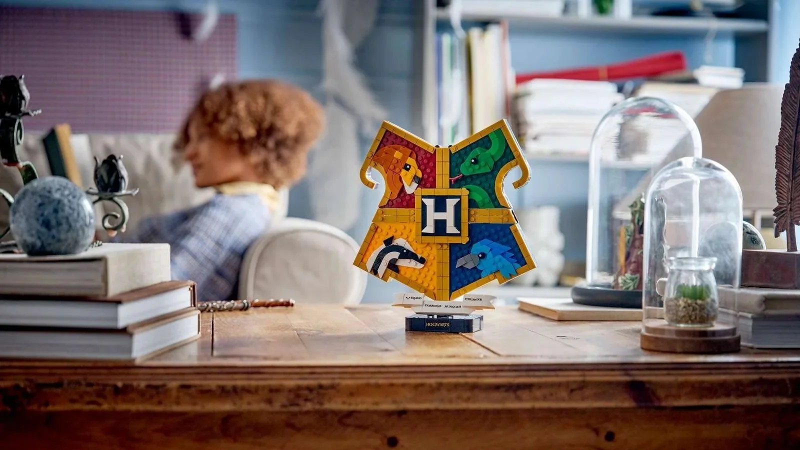 LEGO Hogwarts House Crest set displayed on a wooden desk between stacked books and glass cloches