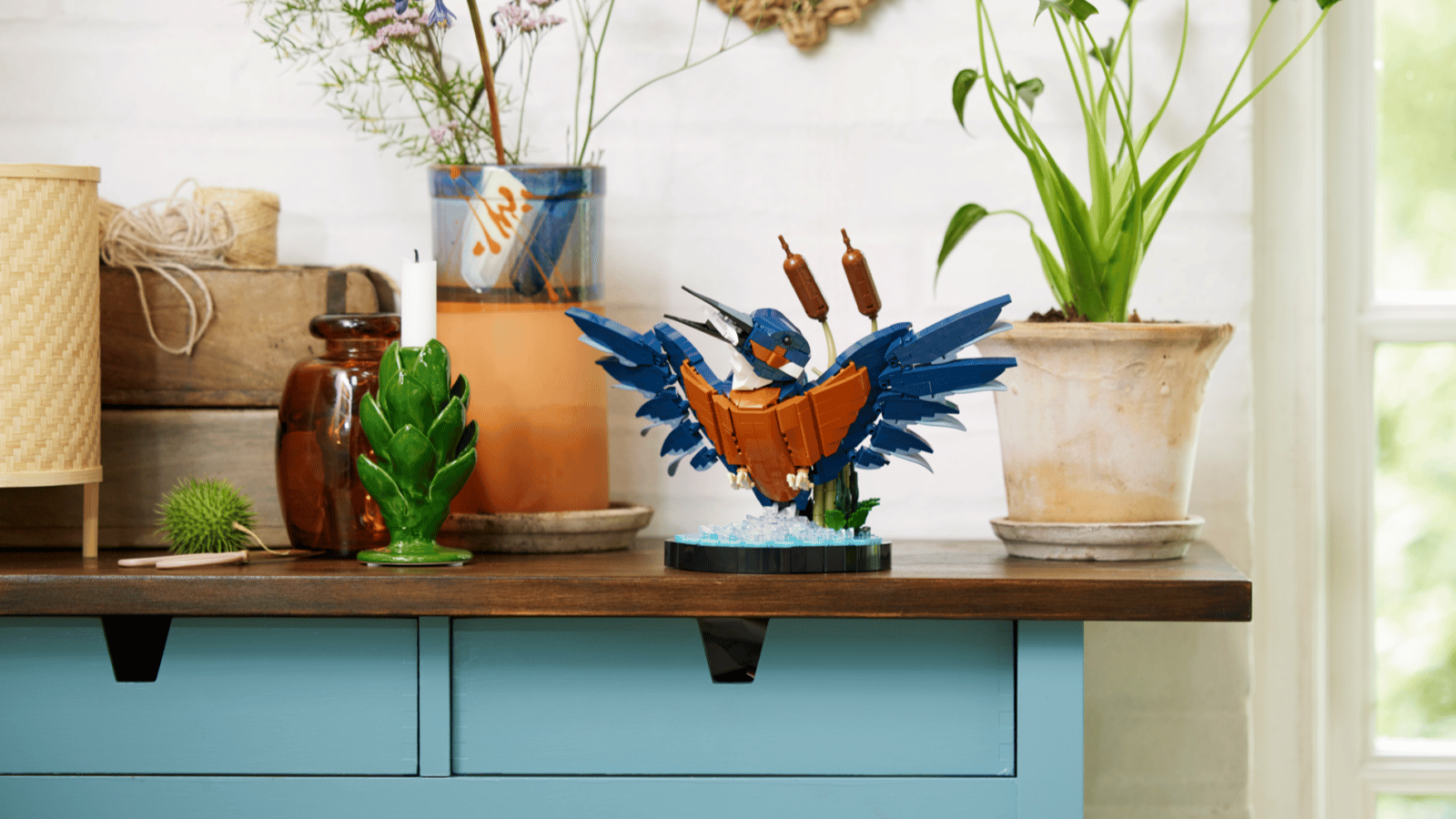 A LEGO Kingfisher Bird sculpture with wings spread sits on a wooden table above a blue drawer unit, surrounded by fresh flowers and a potted