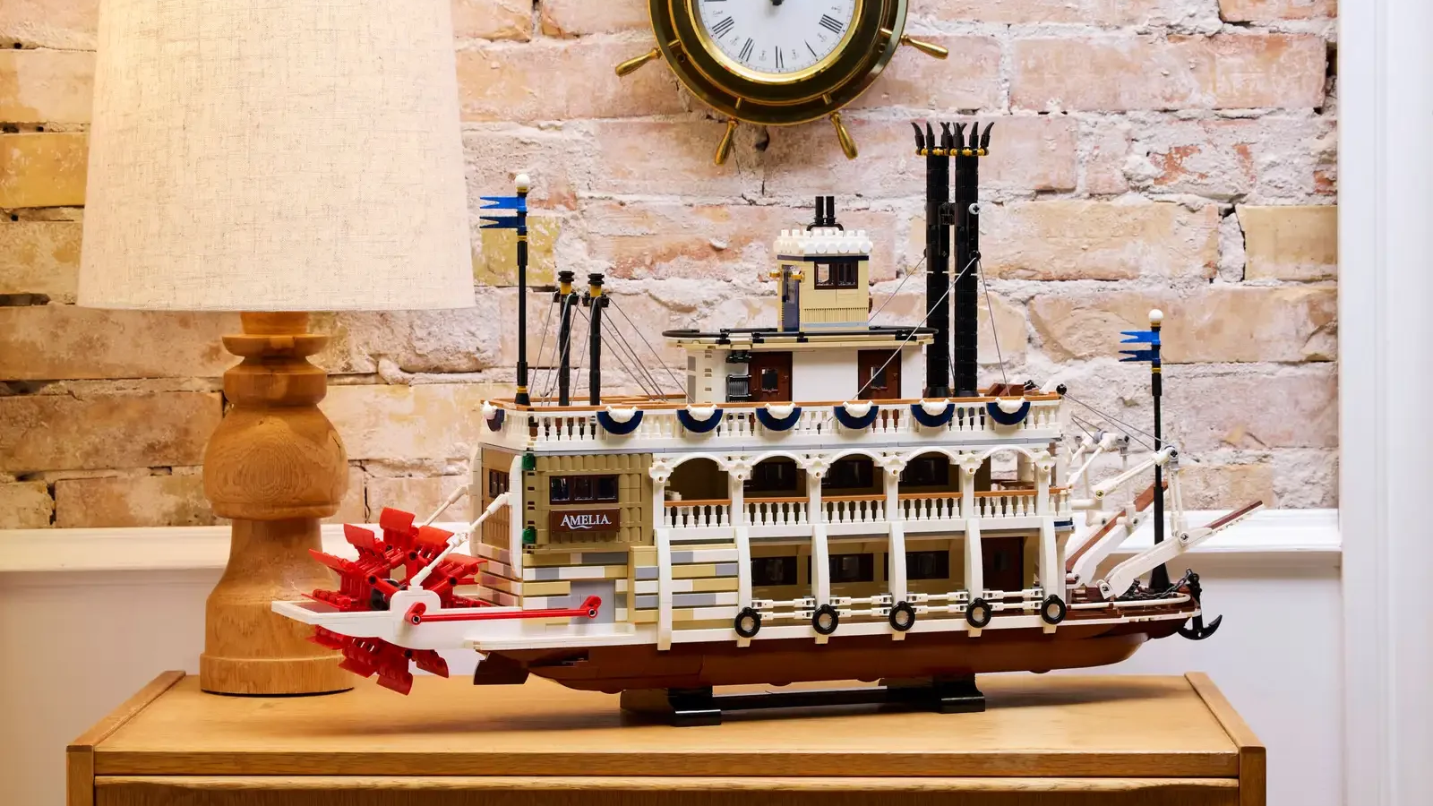 LEGO River Steamboat model named Amelia with a red paddle wheel, displayed on a wooden cabinet against a brick wall beneath a ship's wheel clock
