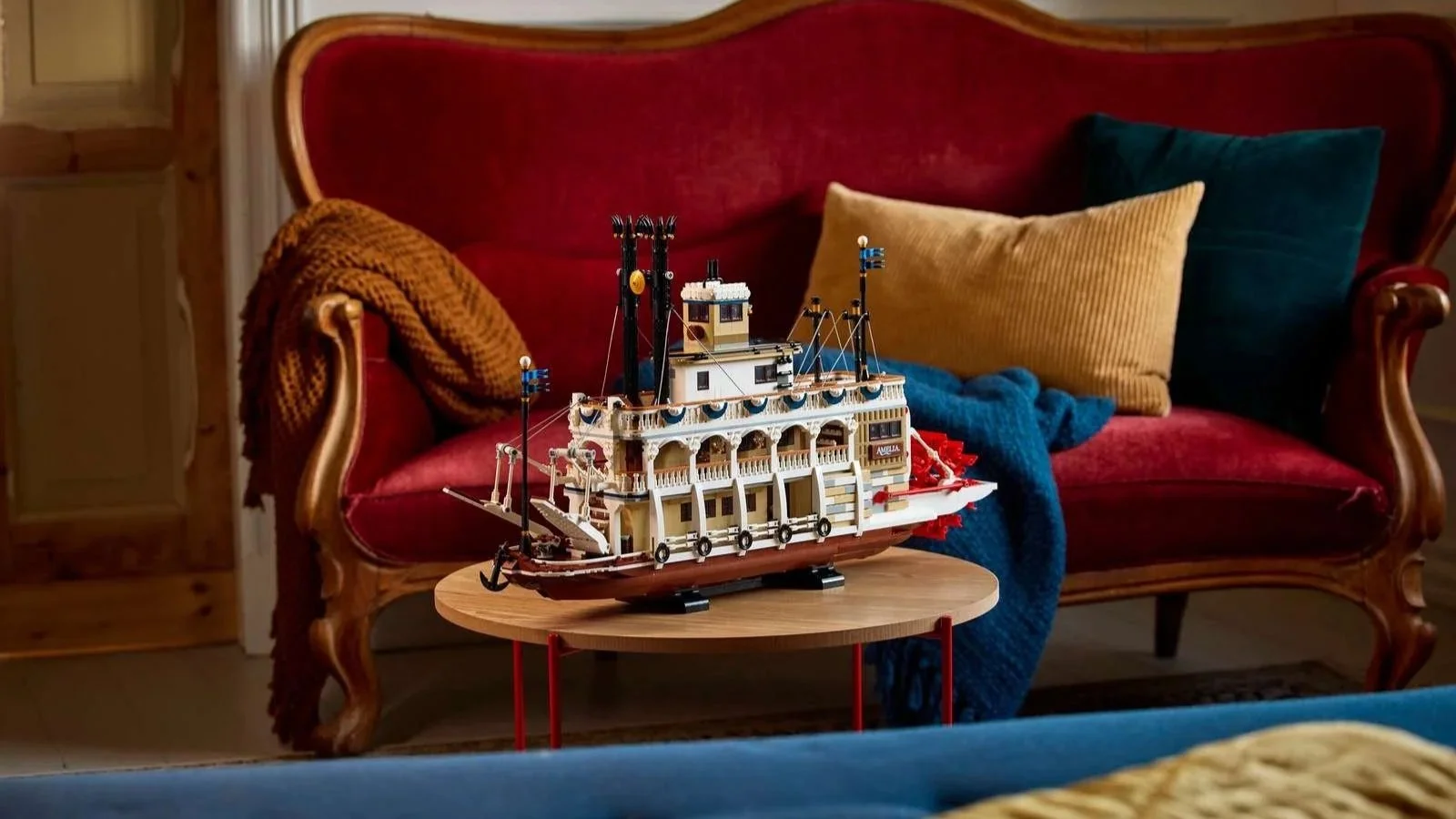 LEGO River Steamboat model named Amelia with a red paddle wheel, displayed on a round coffee table in front of a red velvet sofa with throw pillows