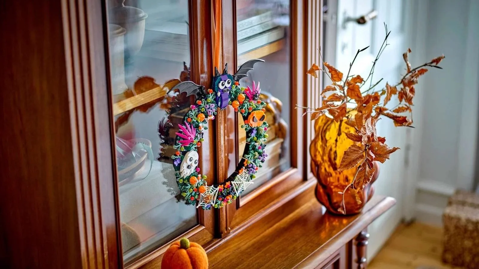 LEGO Halloween Wreath set hanging on a glass-fronted wooden cabinet, decorated with a purple bat, pumpkins, a ghost and spider webs