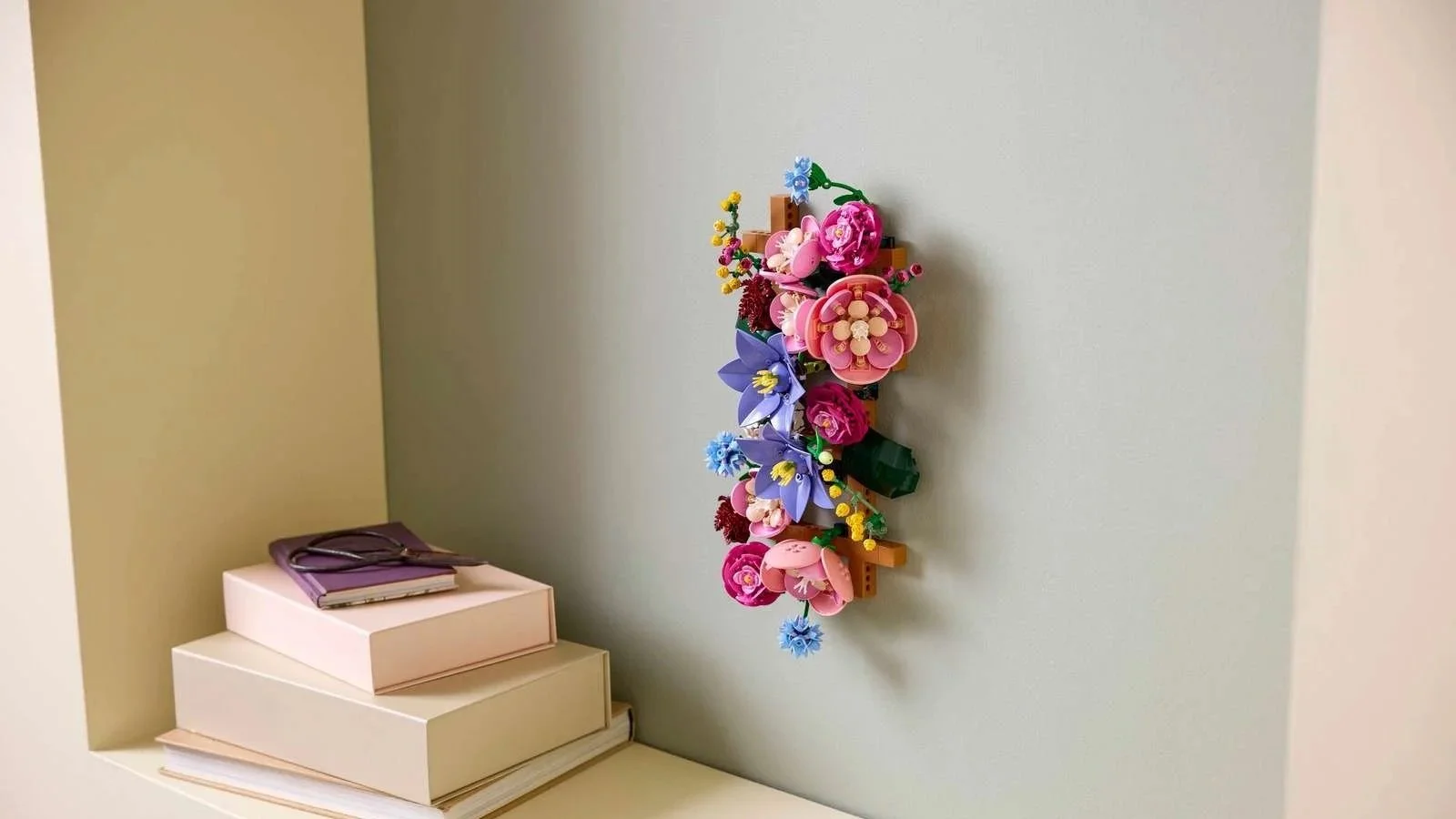 A LEGO Flower Wall arrangement of pink and purple blooms mounted on a wall above a shelf with stacked boxes and a notebook