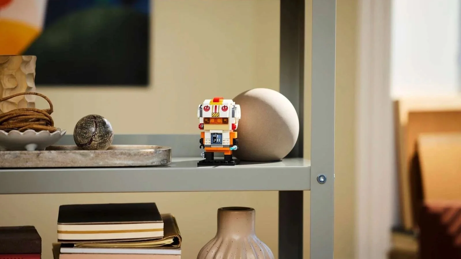 LEGO Luke Skywalker Rebel Pilot buildable figure in an orange and white flight suit and helmet, displayed on a gray shelf beside books and decor
