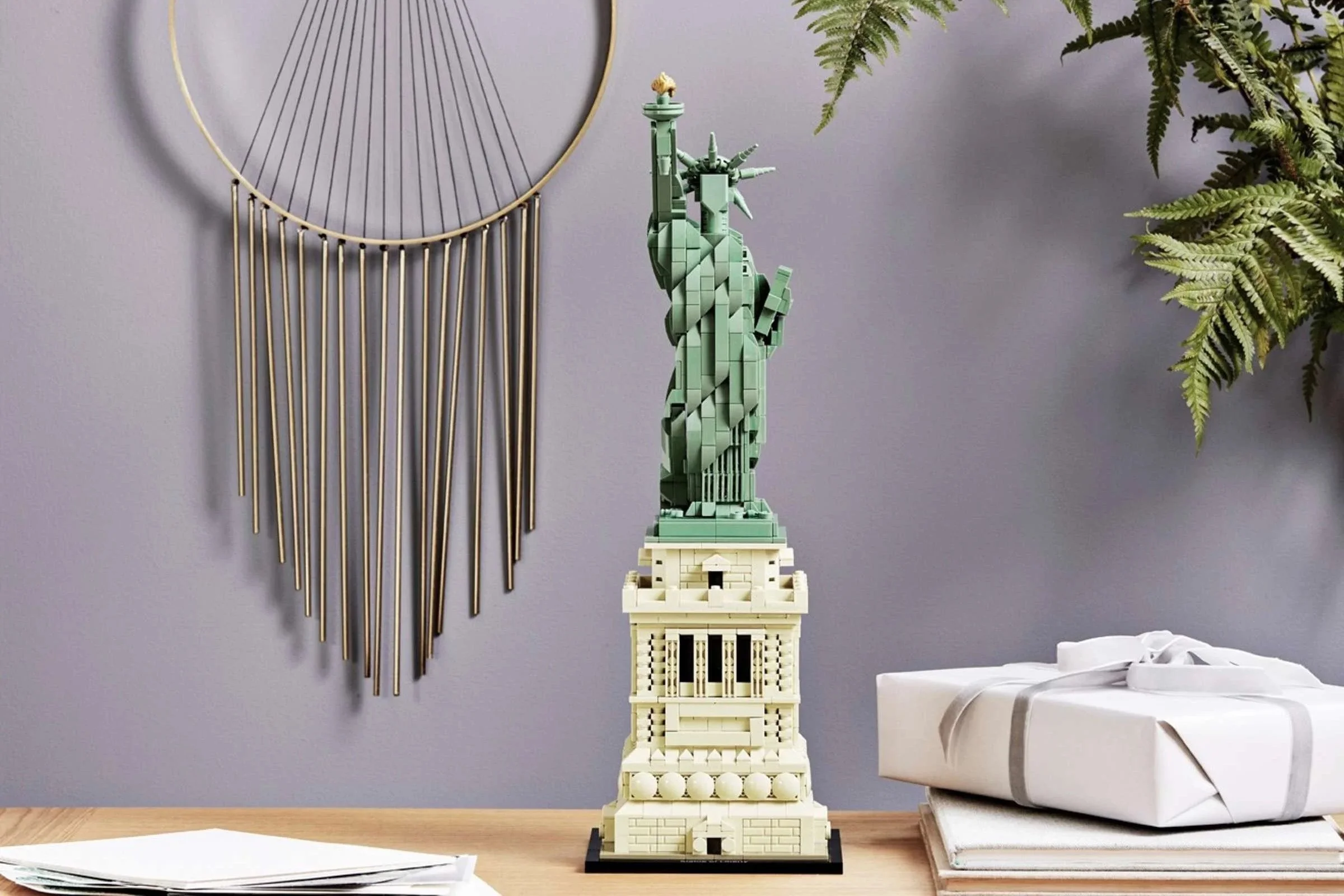 LEGO Statue of Liberty model on a light wood dresser with wrapped gifts, a brass wall hanging, and fern fronds