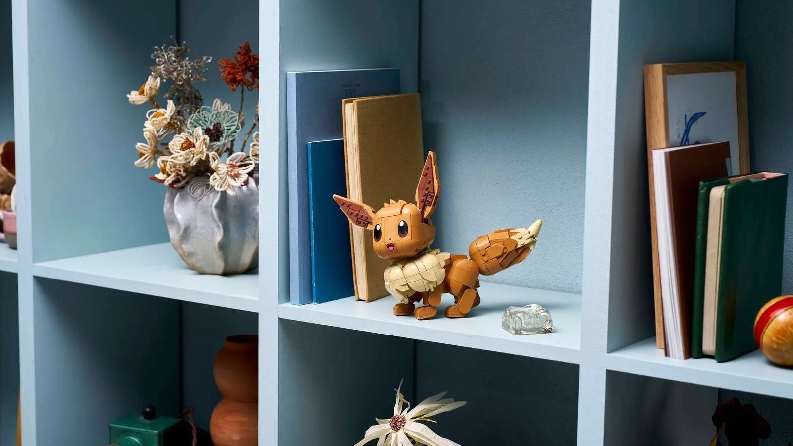 LEGO Eevee set showing the brown Pokémon character with big ears and a fluffy cream collar on a pale blue shelf beside books and dried flowers