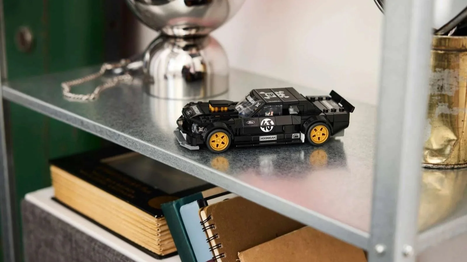 LEGO Ford Mustang Hoonicorn model car in black with Hoonigan branding and gold wheels, displayed on a metal shelf above a stack of books