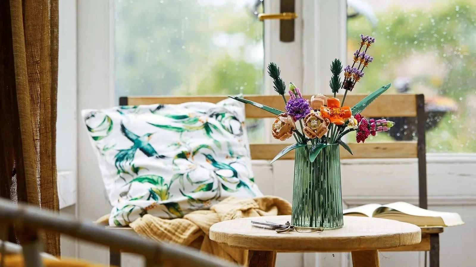 LEGO Flower Bouquet in a ribbed green vase on a round wooden stool beside a bright window seat