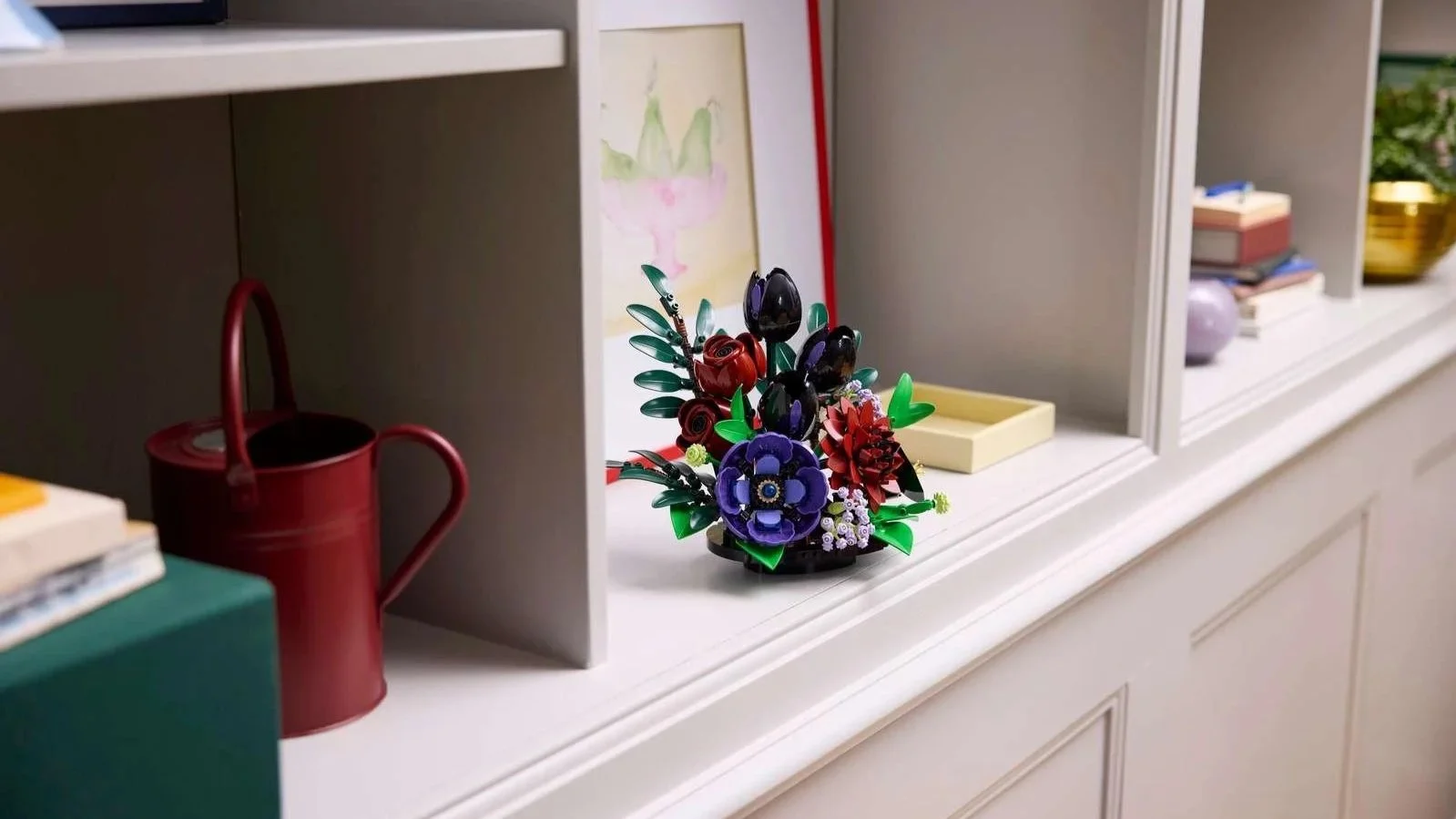A LEGO Dark Flower Arrangement with black tulips and red roses sits on a shelf beside a red watering can and stacked books