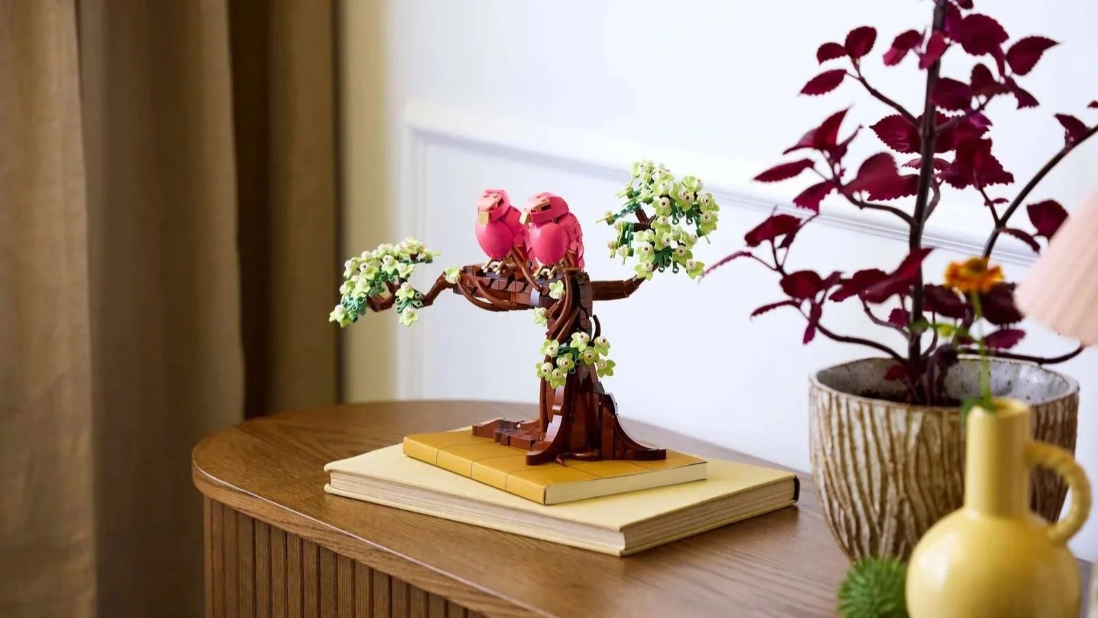 LEGO Love Birds branch sculpture with two pink birds among white blossoms, displayed on stacked books atop a wooden console beside a small potted plant