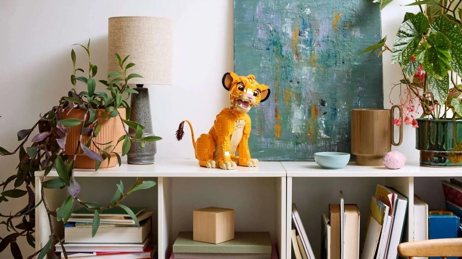 LEGO Young Simba the Lion King set displayed on a white shelf between houseplants and an abstract painting