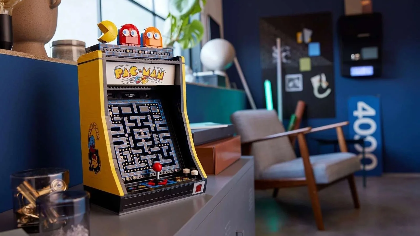 LEGO PAC-MAN Arcade set in yellow with PAC-MAN and ghost figures on top, displayed on a counter beside a potted plant