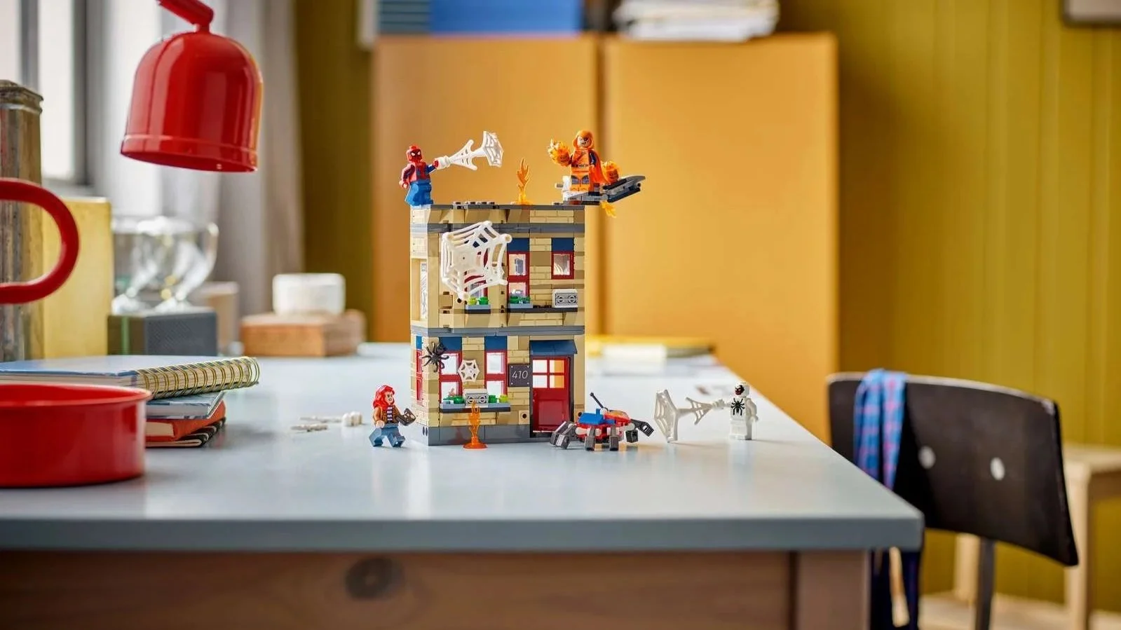 LEGO Peter Parker's Apartment set with Spider-Man shooting webs from the rooftop of a tan brick building on a desk, facing a fiery villain on a glider