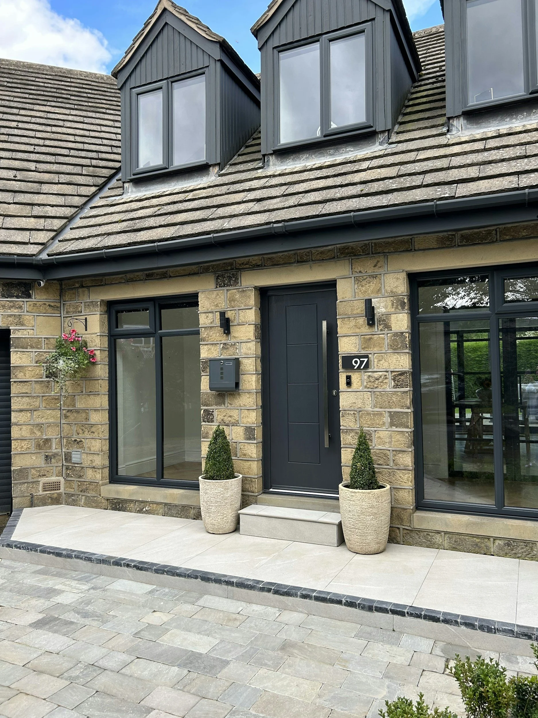 Modern house entrance with black front door, two tall potted green plants, large window, and stone exterior. Two dormer windows on the roof, a hanging flower pot, and a driveway made of gray paving stones.