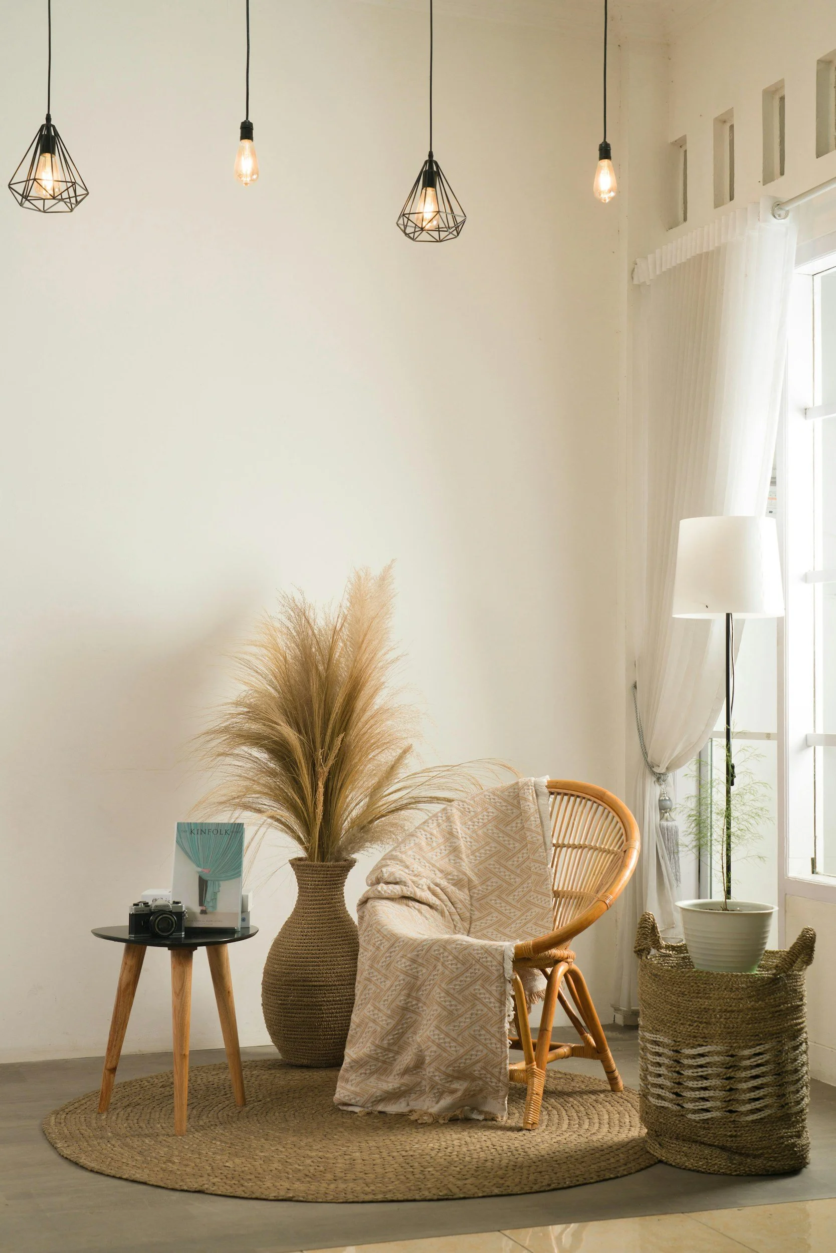 A cozy, minimalistic corner of a living space with a round woven rug, a rattan chair, and a wicker basket. There is a small wooden side table holding a vintage camera, a white book, and a magazine titled 'Kinfolk.' A large ceramic vase with pampas grass is positioned behind the table. In the background, a floor lamp with a white shade stands near a window with sheer white curtains, and a series of Edison-style pendant lights hang from the ceiling.